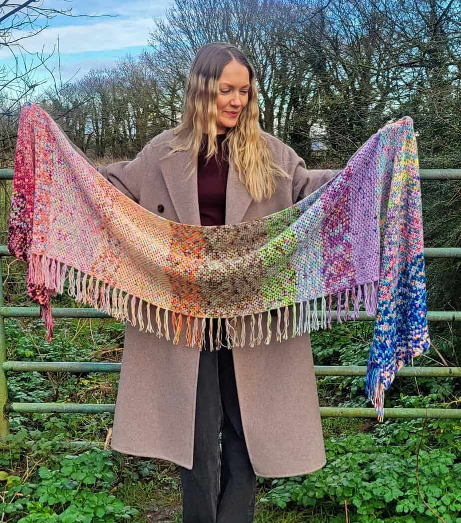 woman holding a long crochet shawl made with mini skeins standing infront of an old gate