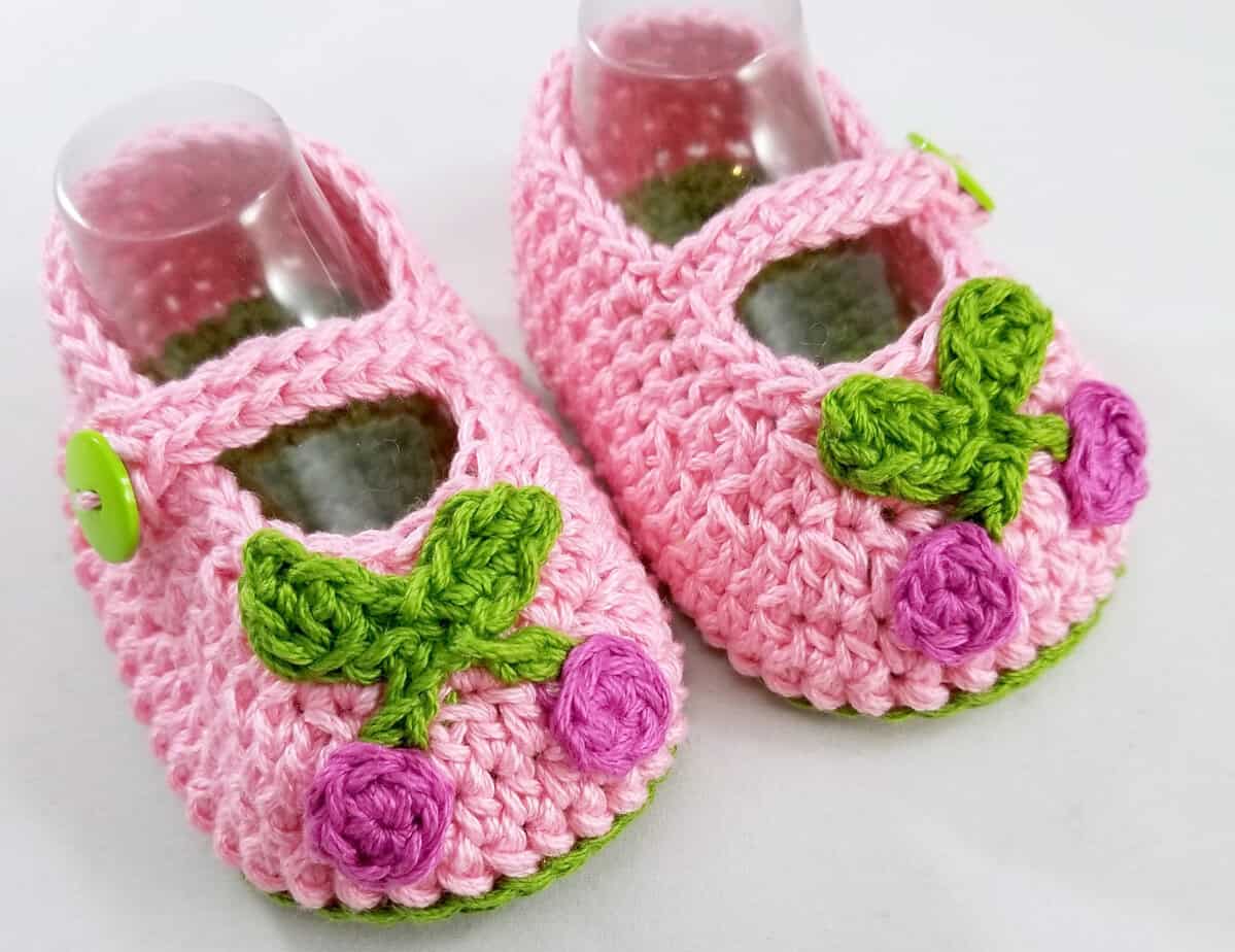 Pink crochet baby shoes with green and purple floral embellishments.